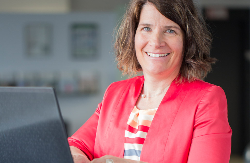 Susanne Gensinger sitzt am Schreibtisch, lächelt den Betrachter an und macht sich Notizen zur Gesundheitskommunikation für Ihr Unternehmen